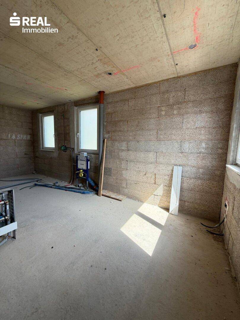 Unfertiges Badezimmer im Rohbauzustand mit sichtbaren Installationen und Fenster.