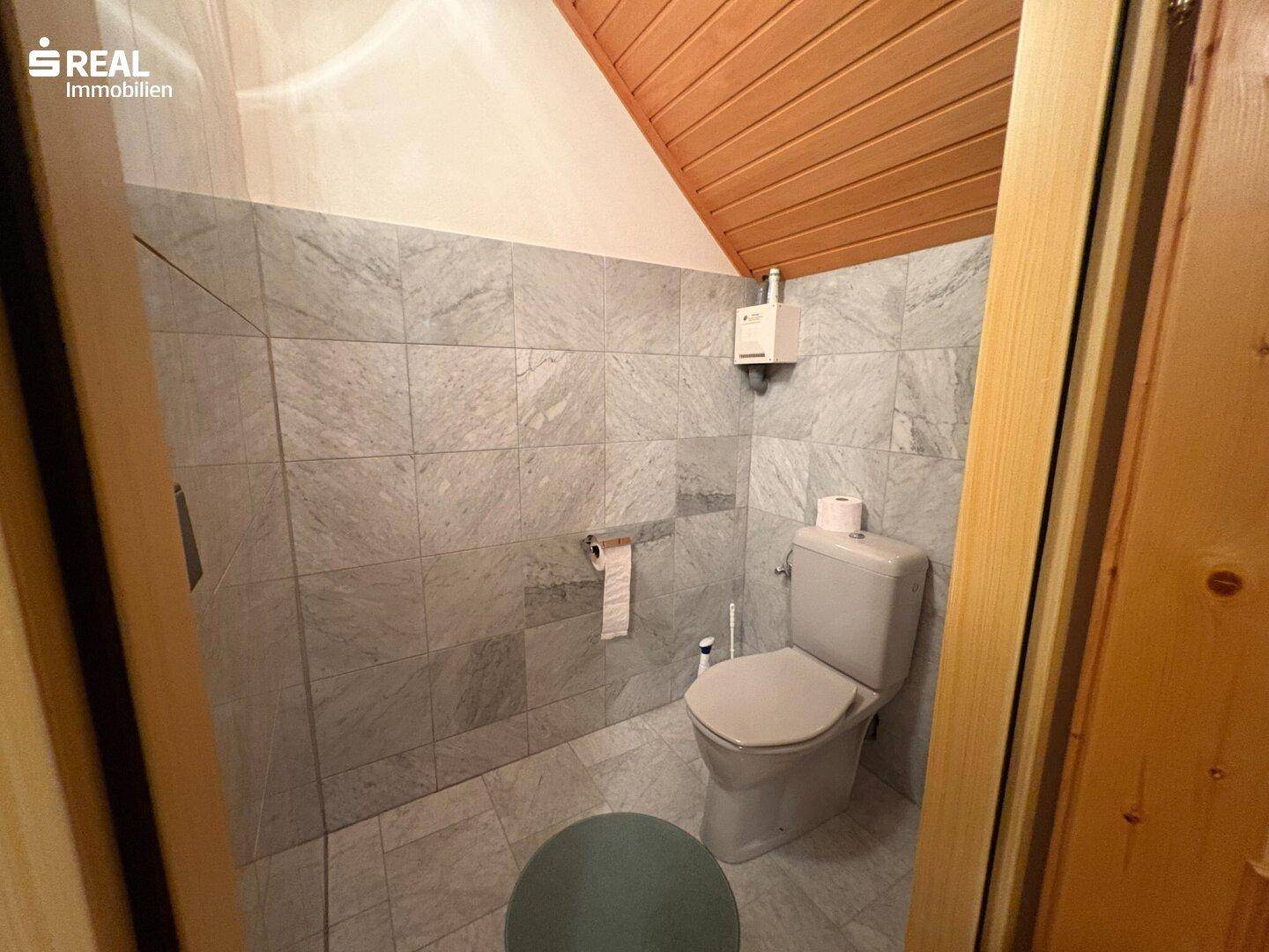 Kleines Gäste-WC mit grauen Fliesen, Toilette und Holzdecke unter Dachschräge.