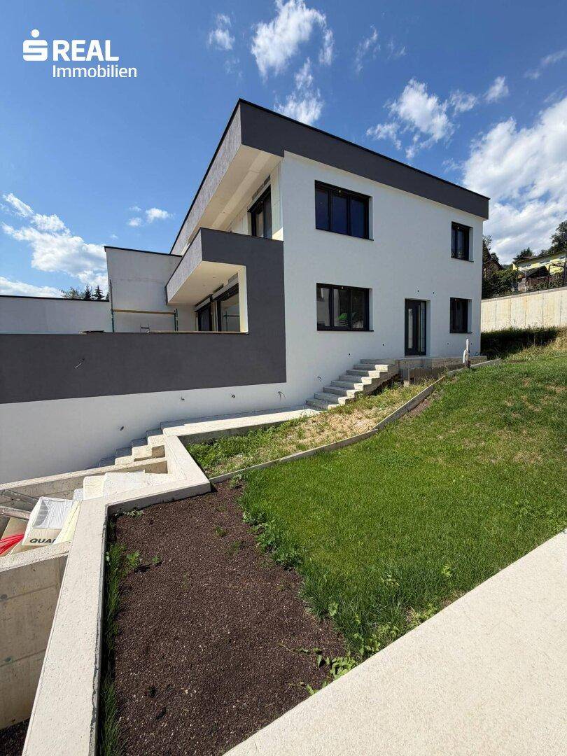 Moderne Hausfassade mit weißem Putz und dunklen Fenstern, umgeben von einer Grünfläche.