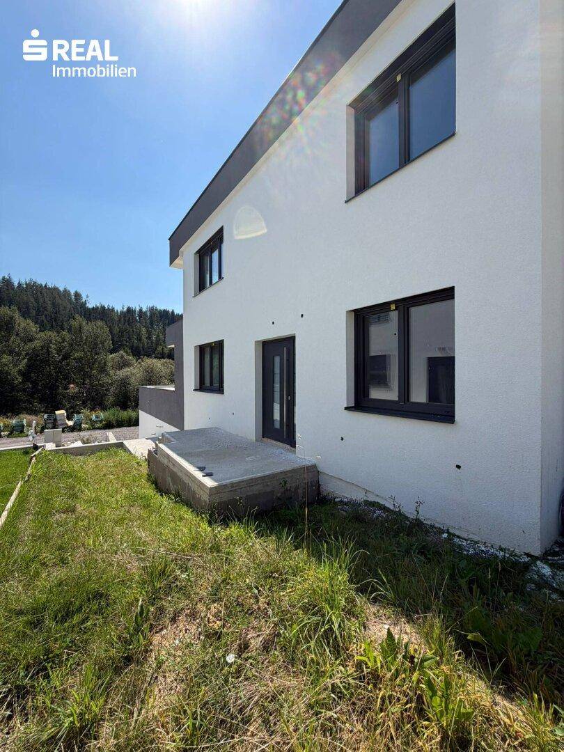 Moderne Hausfassade mit weißem Putz und dunklen Fenstern, umgeben von einer Grünfläche.