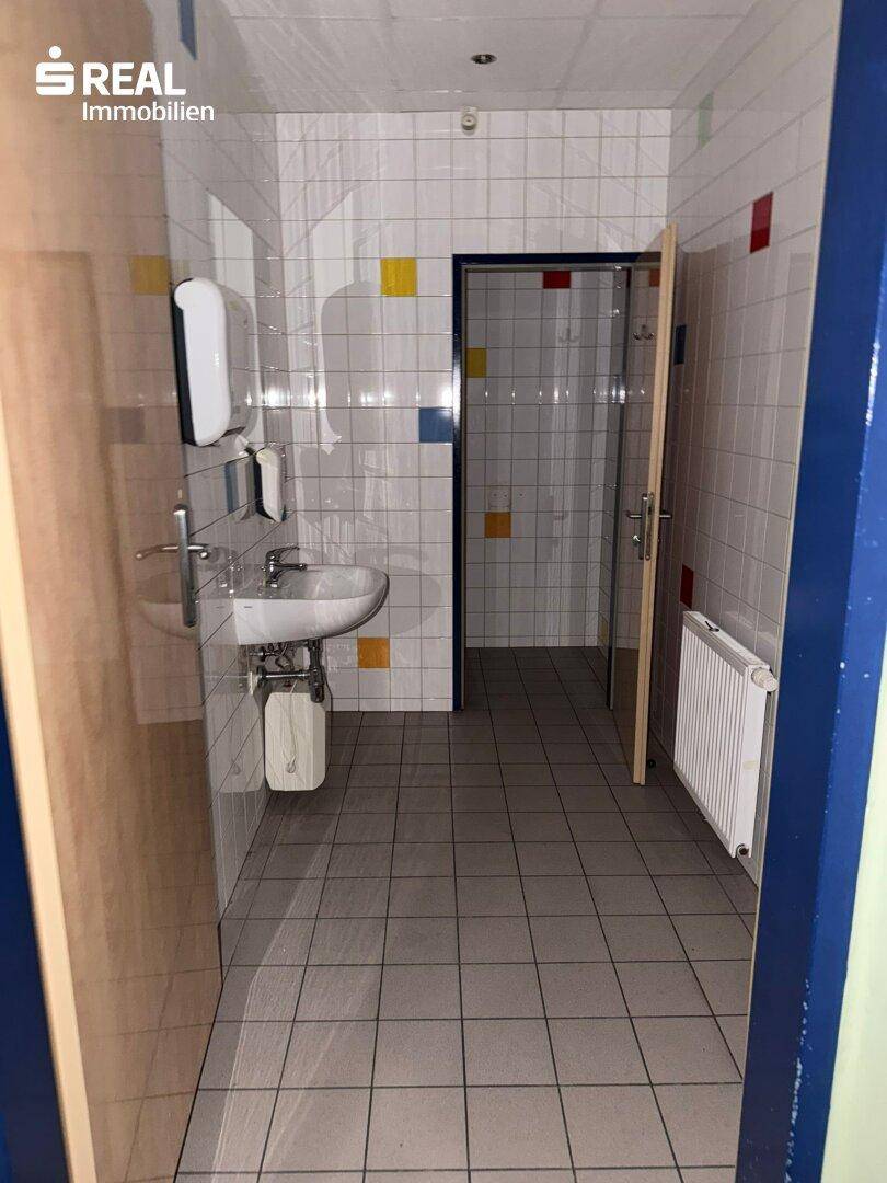 Öffentlicher Toilettenraum mit Waschbecken, Heizkörper und bunten Fliesen an Wänden und Boden.