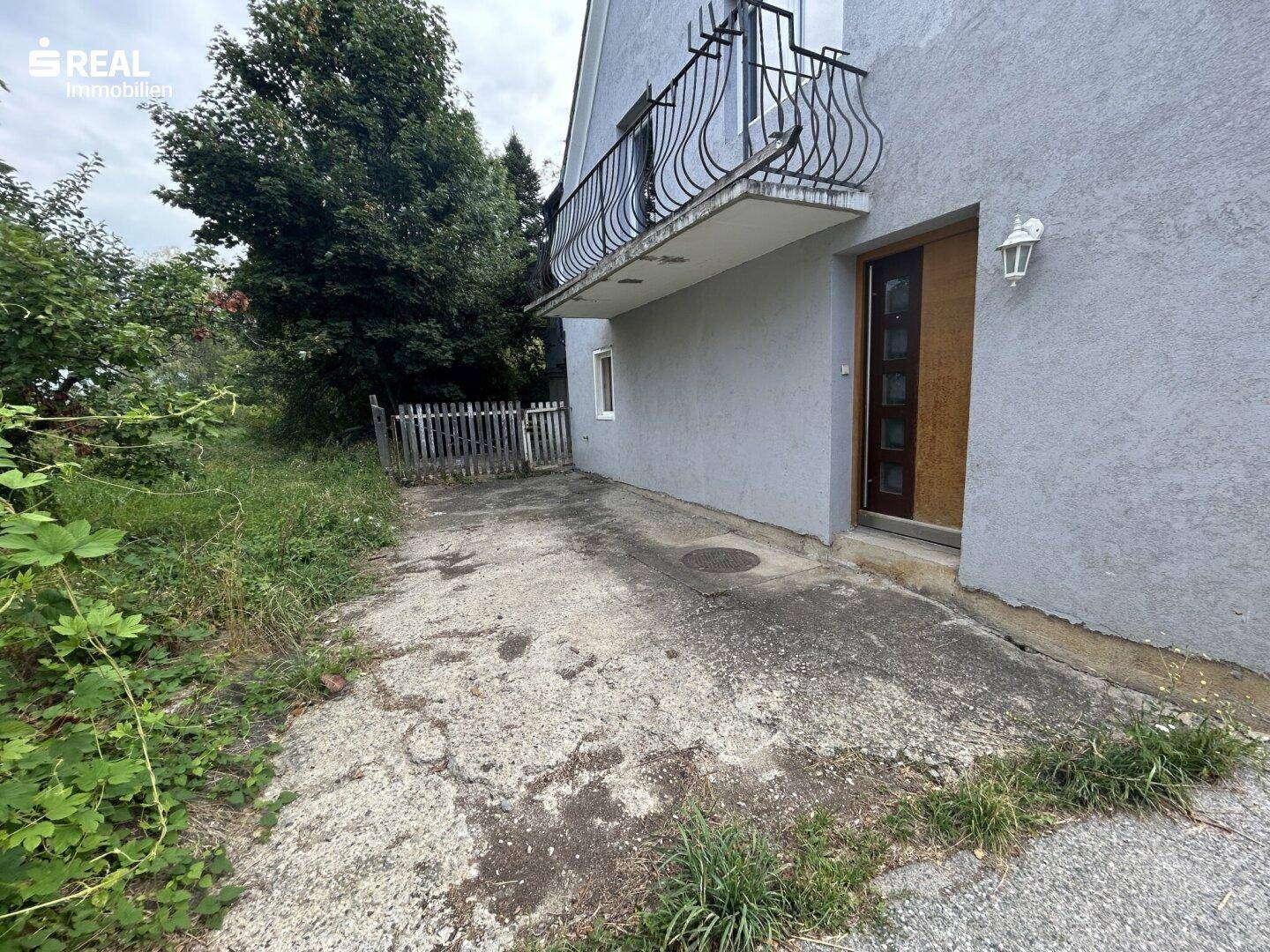 Graue Hausfassade mit Balkon und einem ungepflegten Betonweg im Vordergrund.