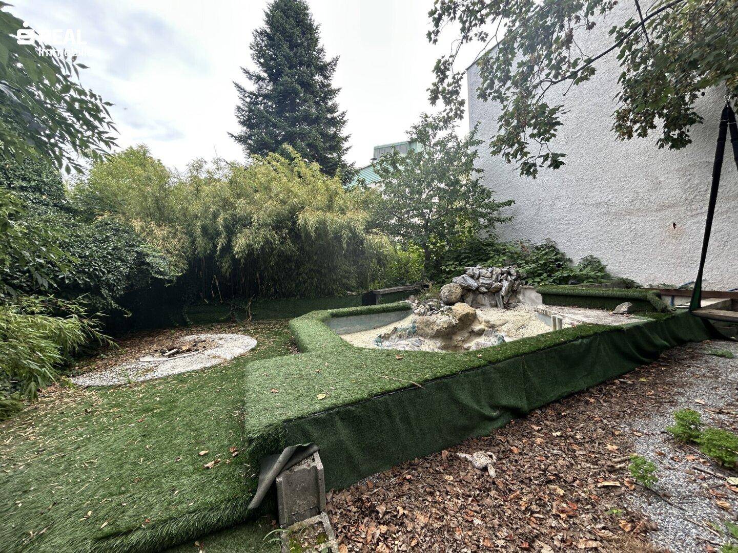 Gartenbereich mit Kunstrasen, einem kleinen Teich und üppiger Bepflanzung.