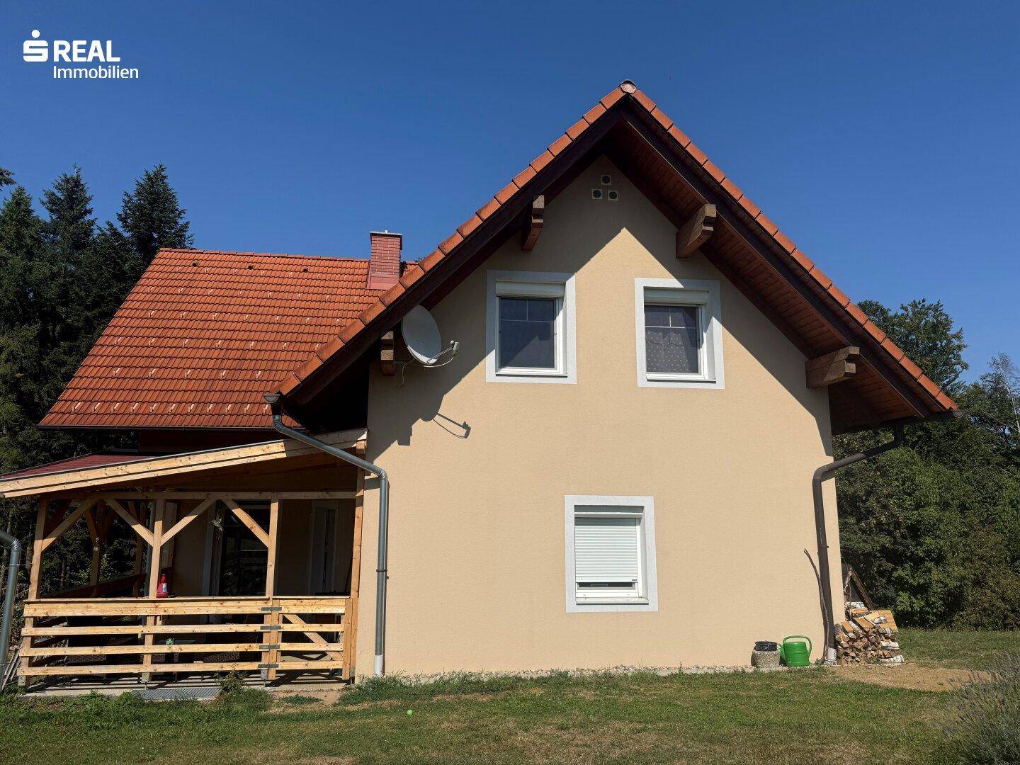Hausfassade mit Satteldach, beiger Putzfassade und überdachtem Holzvorbau in ländlicher Umgebung.