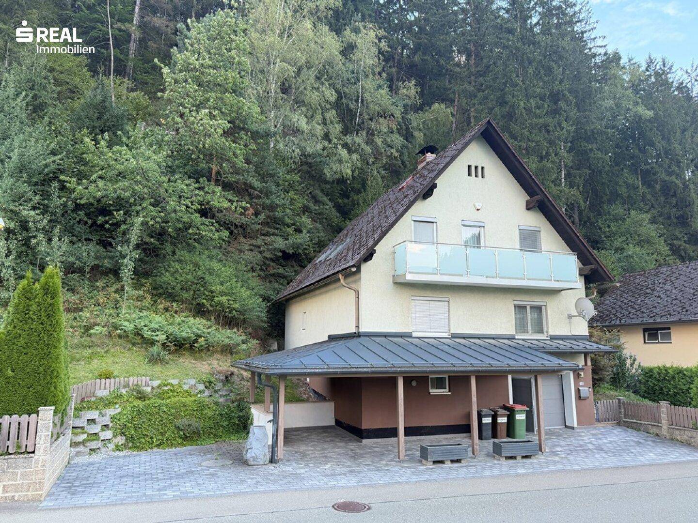 Seitenansicht des Hauses mit Carport und Balkon, eingebettet in eine grüne Hügellandschaft.