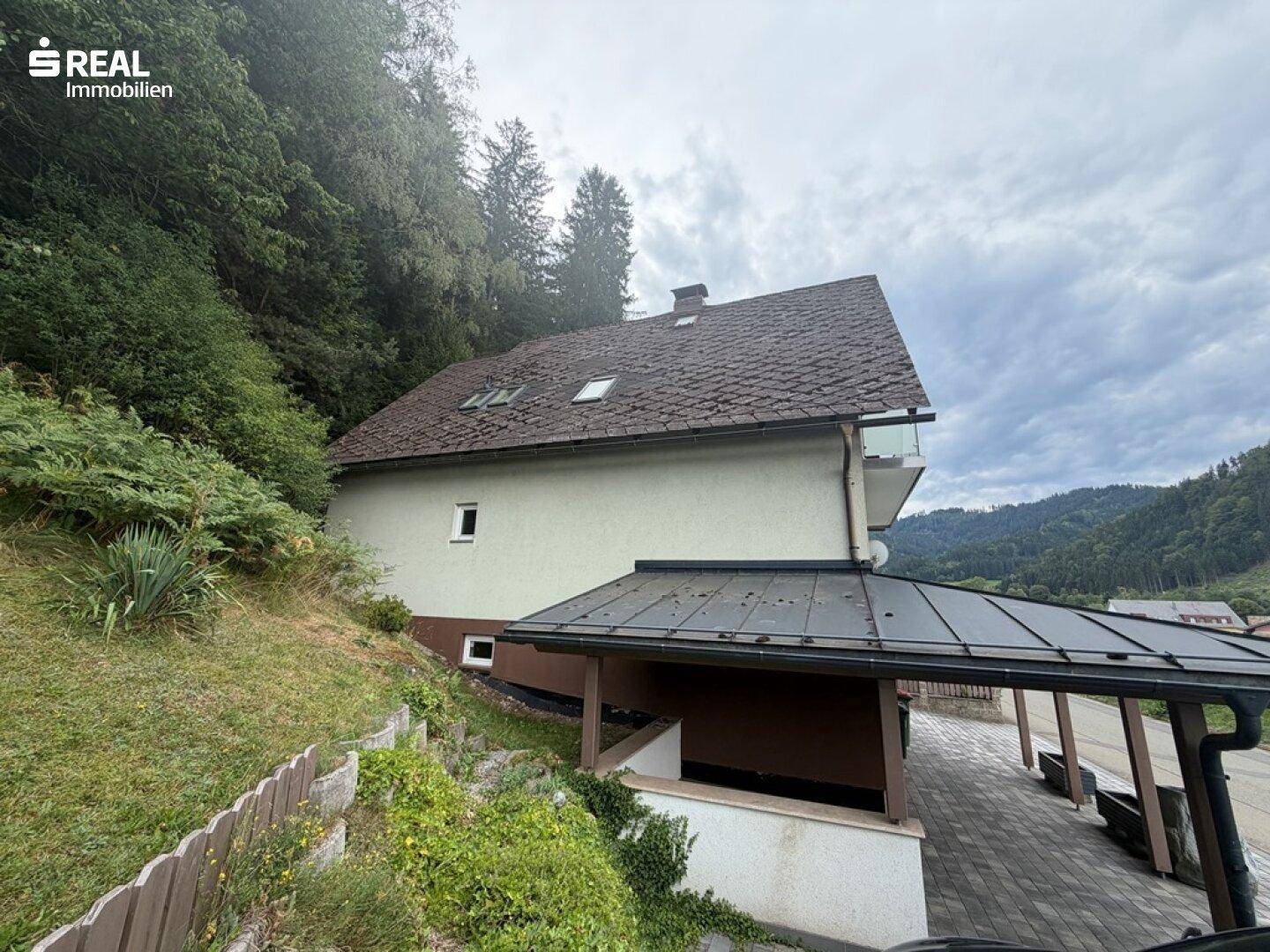 Seitenansicht des Hauses mit Dachfenstern und dem angrenzenden bewaldeten Hang.
