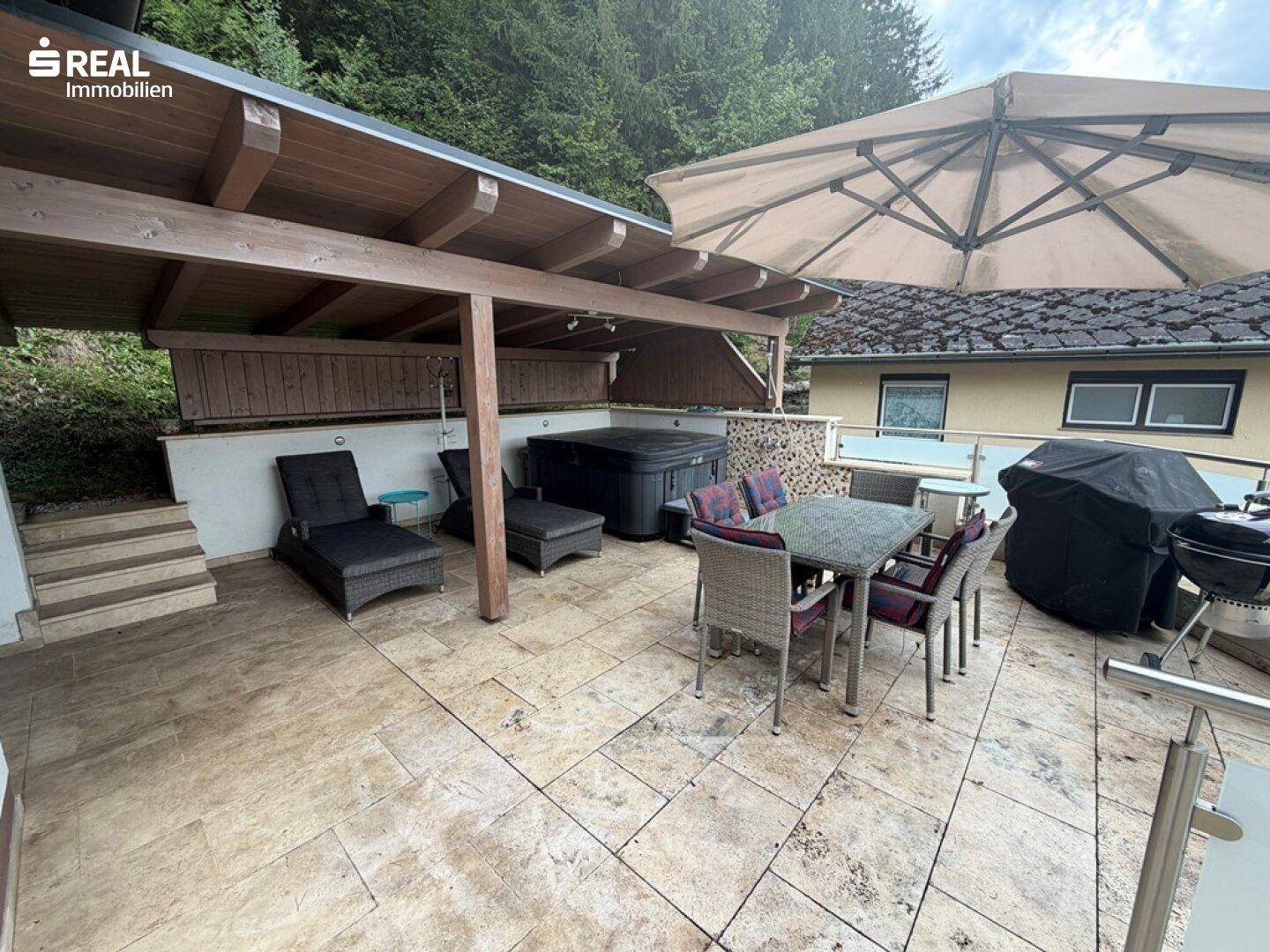 Großzügige Terrasse mit Essbereich, Liegestühlen, Whirlpool und Überdachung, umgeben von Natur.