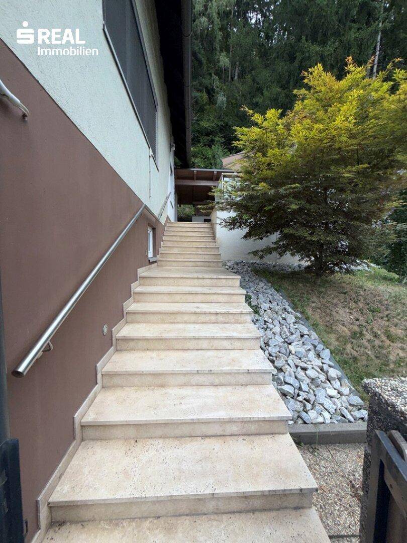 Außentreppe aus hellem Stein führt zum Haus, flankiert von einem Steingarten und Bäumen.