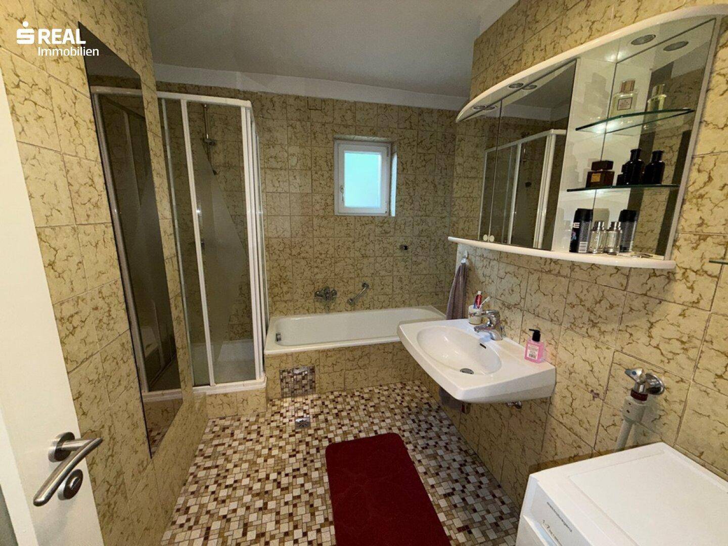 Badezimmer mit gemusterten Fliesen, Badewanne mit Duschabtrennung und Fenster.