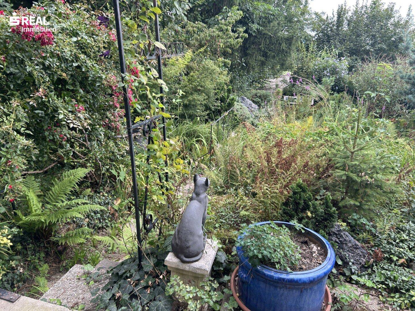 Dekorativer Garten mit einer Katzenstatue, blühenden Pflanzen und einem großen Topf.