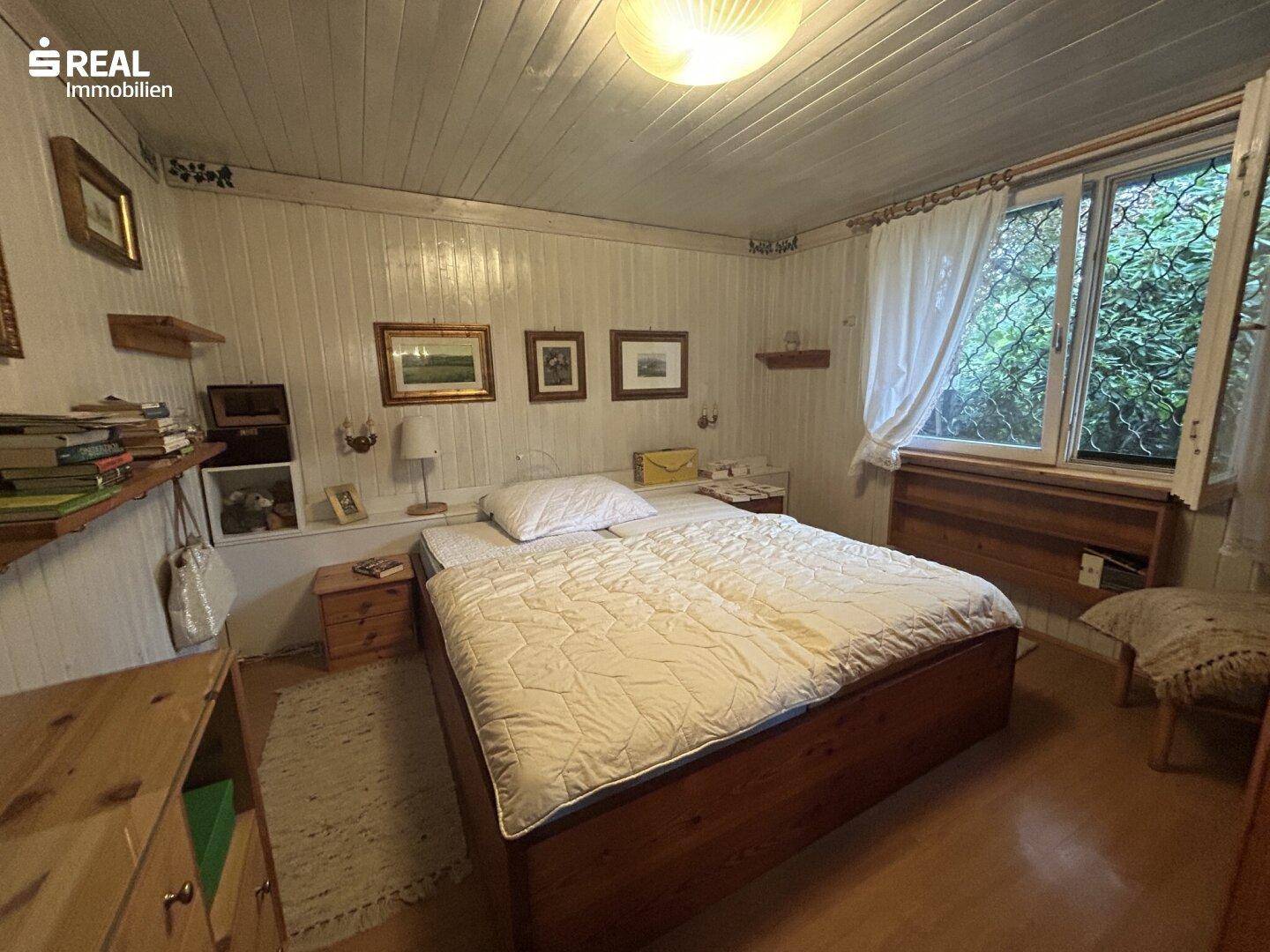 Helles Schlafzimmer mit Holzbett, weiß getäfelten Wänden und Blick ins Grüne.