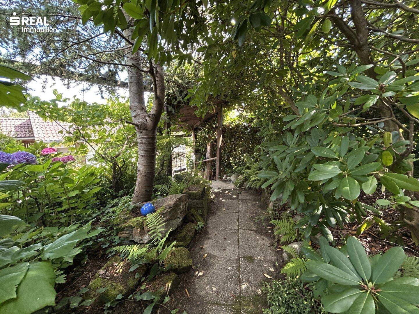 Verwunschener Gartenweg, umgeben von Bäumen und Sträuchern, führt durch das Grüne.