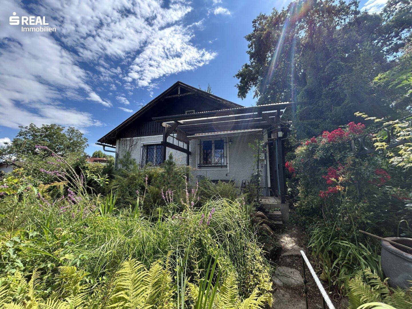 Hausfassade umgeben von wilder Natur und einem sonnigen Gartenweg unter blauem Himmel.