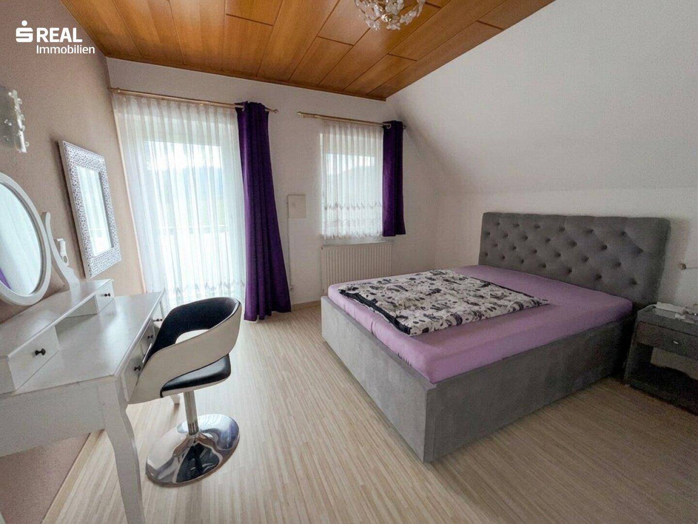 Schlafzimmer mit gepolstertem Bett, Schminktisch und Holzdecke, beleuchtet durch große Fenster.