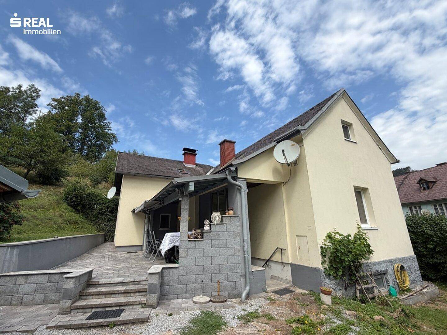 Seitenansicht des Hauses mit gepflasterter Terrasse, Treppenaufgang und Blick auf die grüne Umgebung.