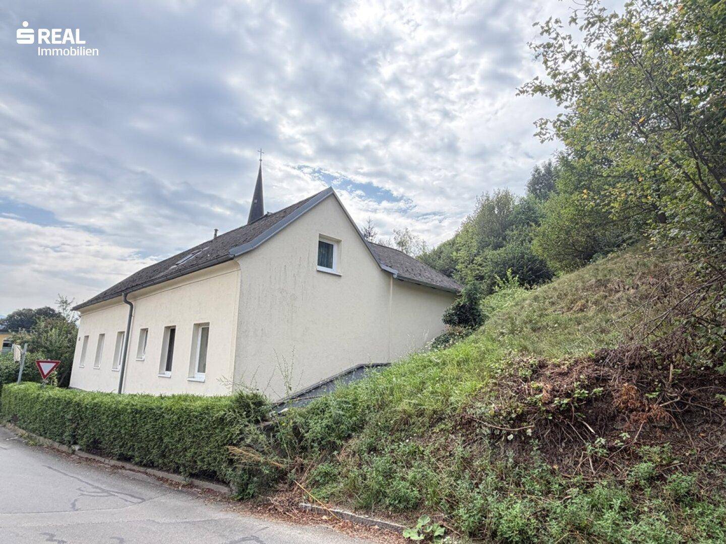 Seitenansicht des Hauses mit schrägem Dach und angrenzendem bewaldeten Hang.