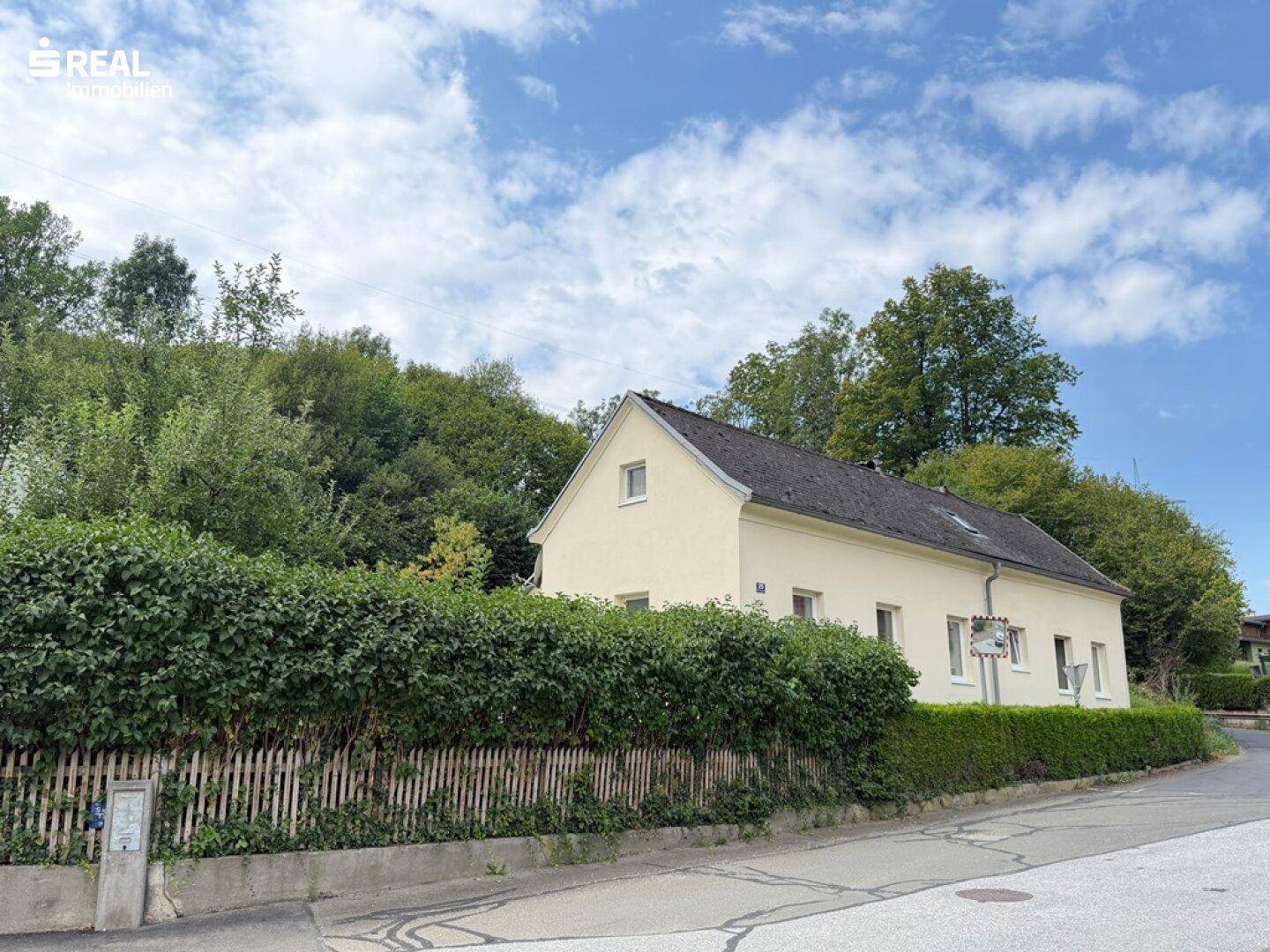 Das Haus von der Straße aus gesehen, umgeben von einer hohen Hecke und Bäumen.