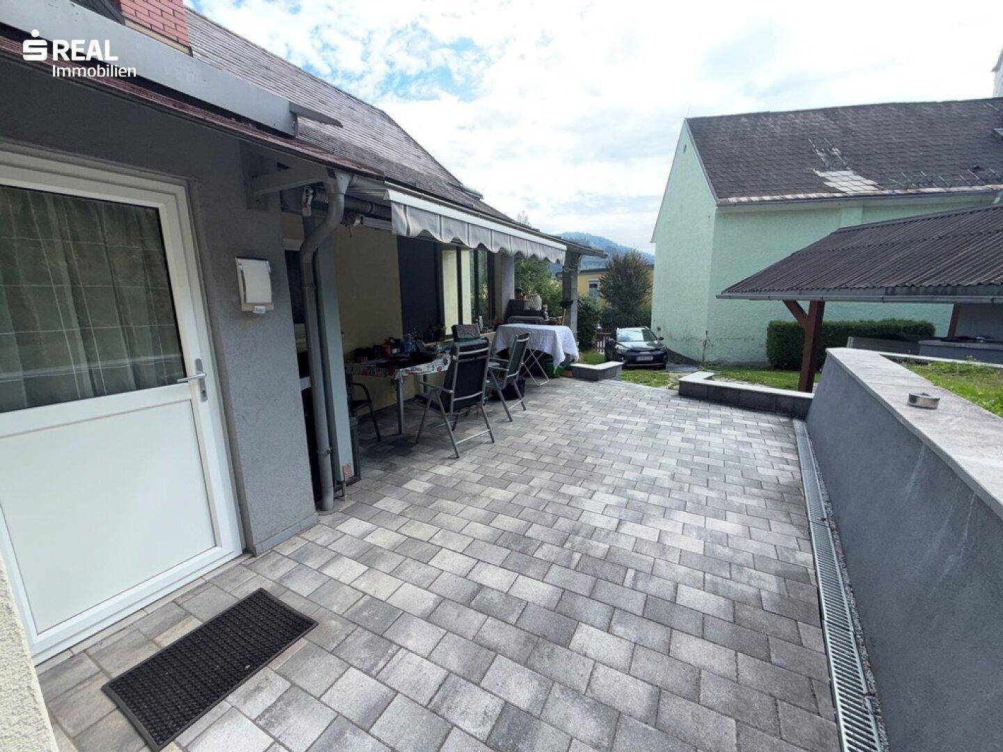Blick von der überdachten Terrasse auf den gepflasterten Hofbereich und die angrenzende Grünfläche.