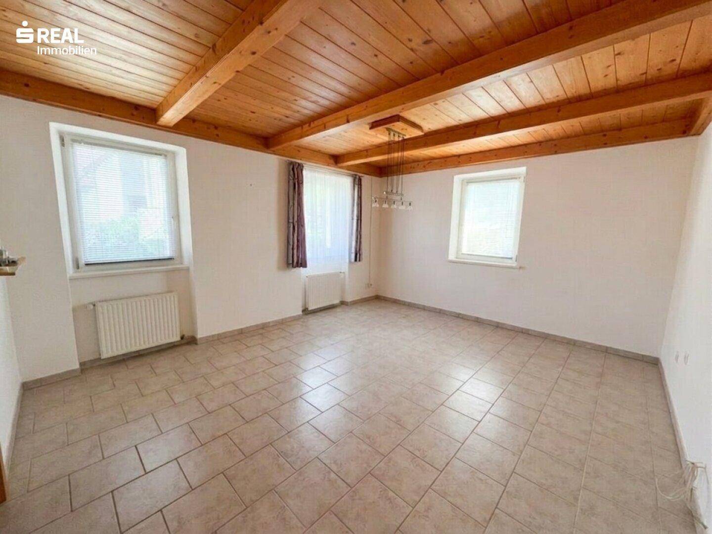 Geräumiger Raum mit Fliesenboden, sichtbaren Holzdeckenbalken und mehreren Fenstern.