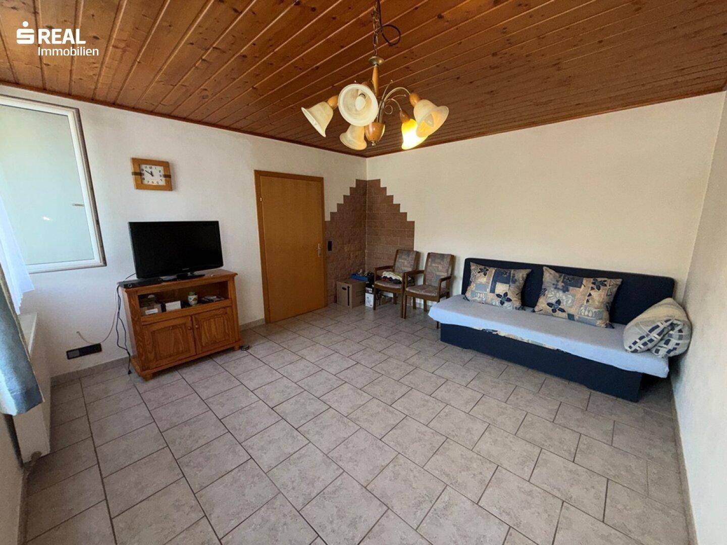 Gemütliches Wohnzimmer mit Fliesenboden, Holzdecke und einfacher Möblierung.