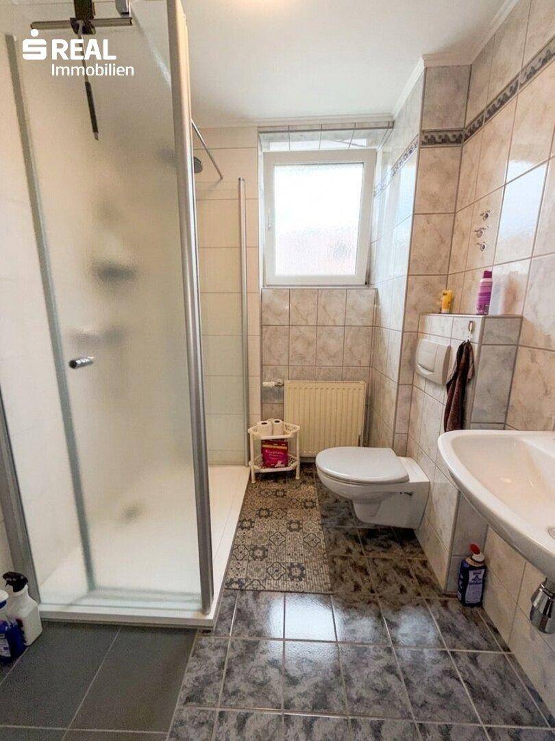 Modernes Badezimmer mit Duschkabine, WC, Waschbecken und Fenster, gefliest in hellen und dunklen Tönen.
