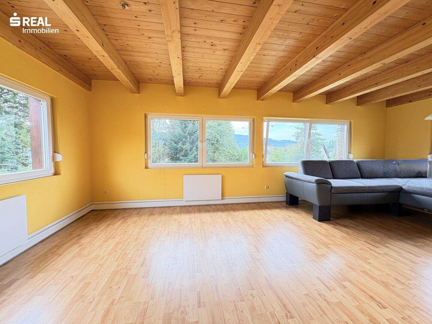 Helles Wohnzimmer mit Holzbalkendecke, gelben Wänden und weitem Blick ins Grüne.