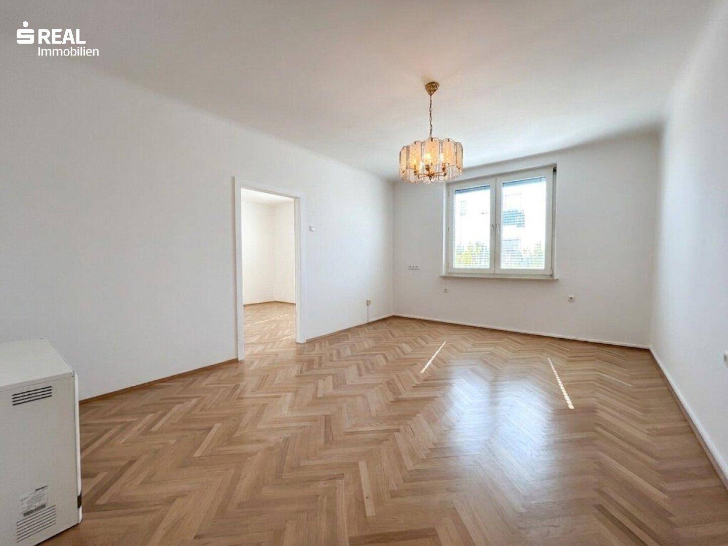 Geräumiges Zimmer mit Fischgrätparkett, großen Fenstern und einem eleganten Kronleuchter, lichtdurchflutet.