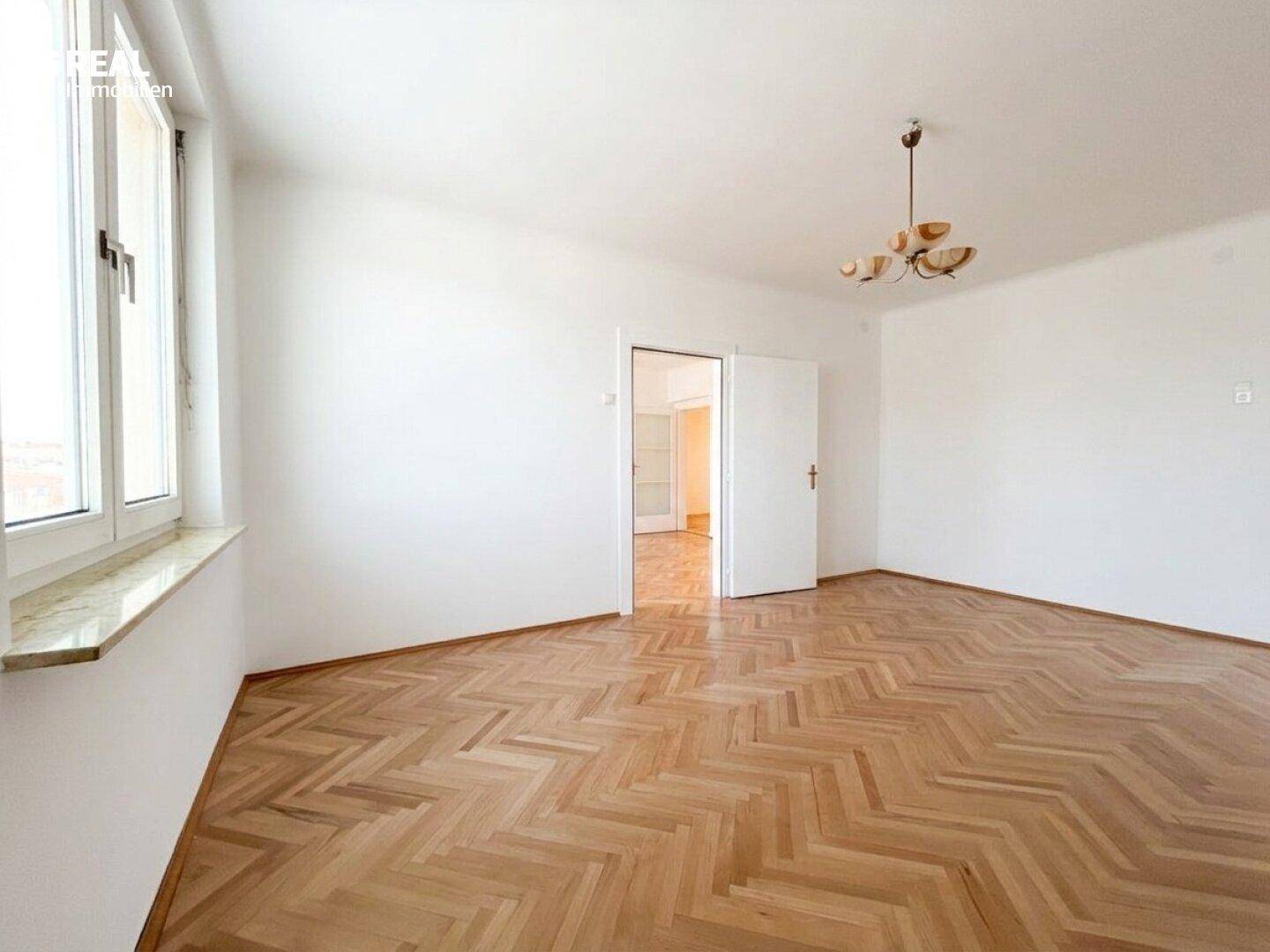 Lichtdurchflutetes Zimmer mit klassischem Fischgrätparkett und großen Fenstern, die viel Tageslicht hereinlassen.
