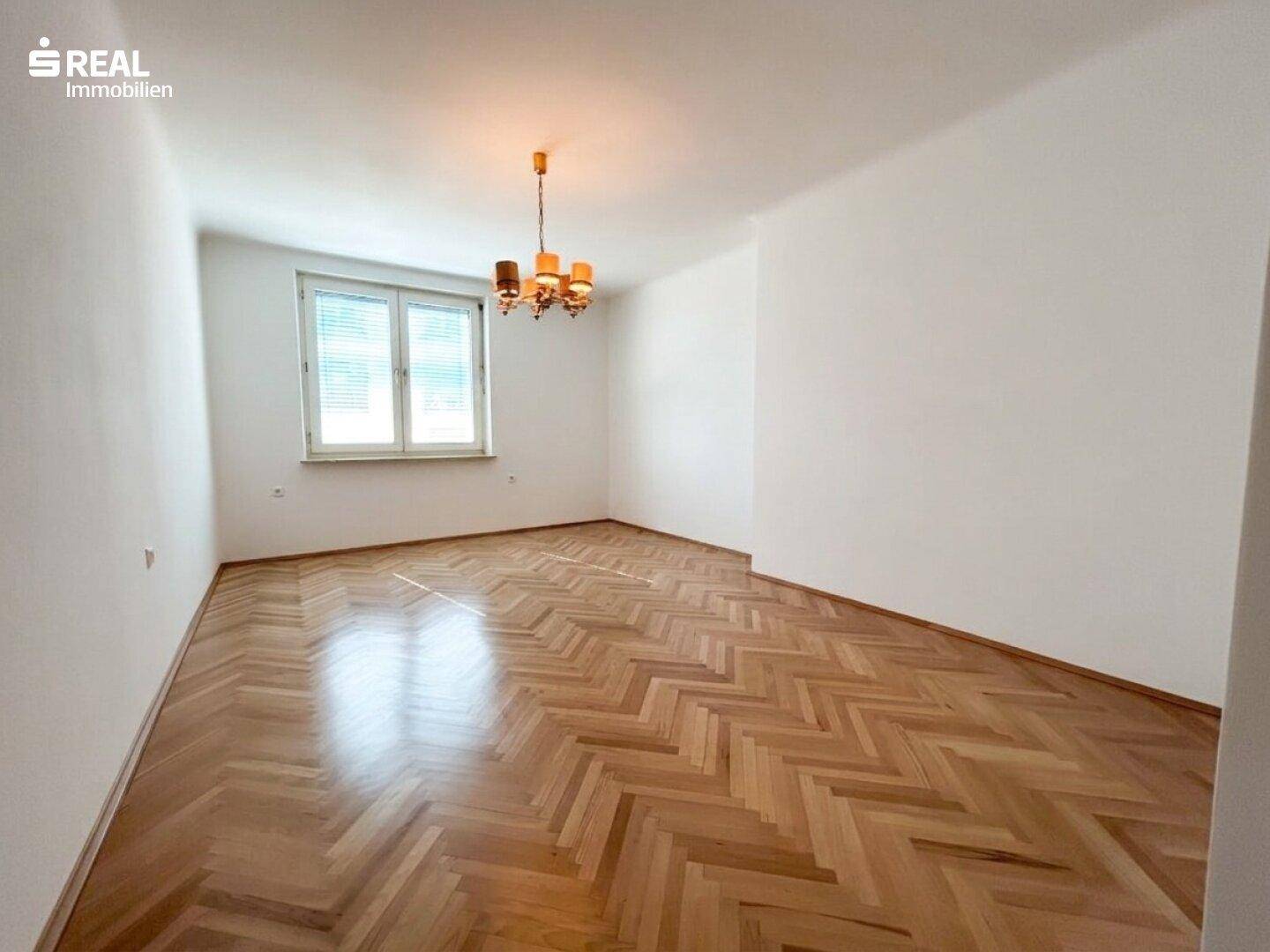 Helles Zimmer mit Fischgrätparkettboden und einem Fenster mit Jalousien, das viel Tageslicht bietet.