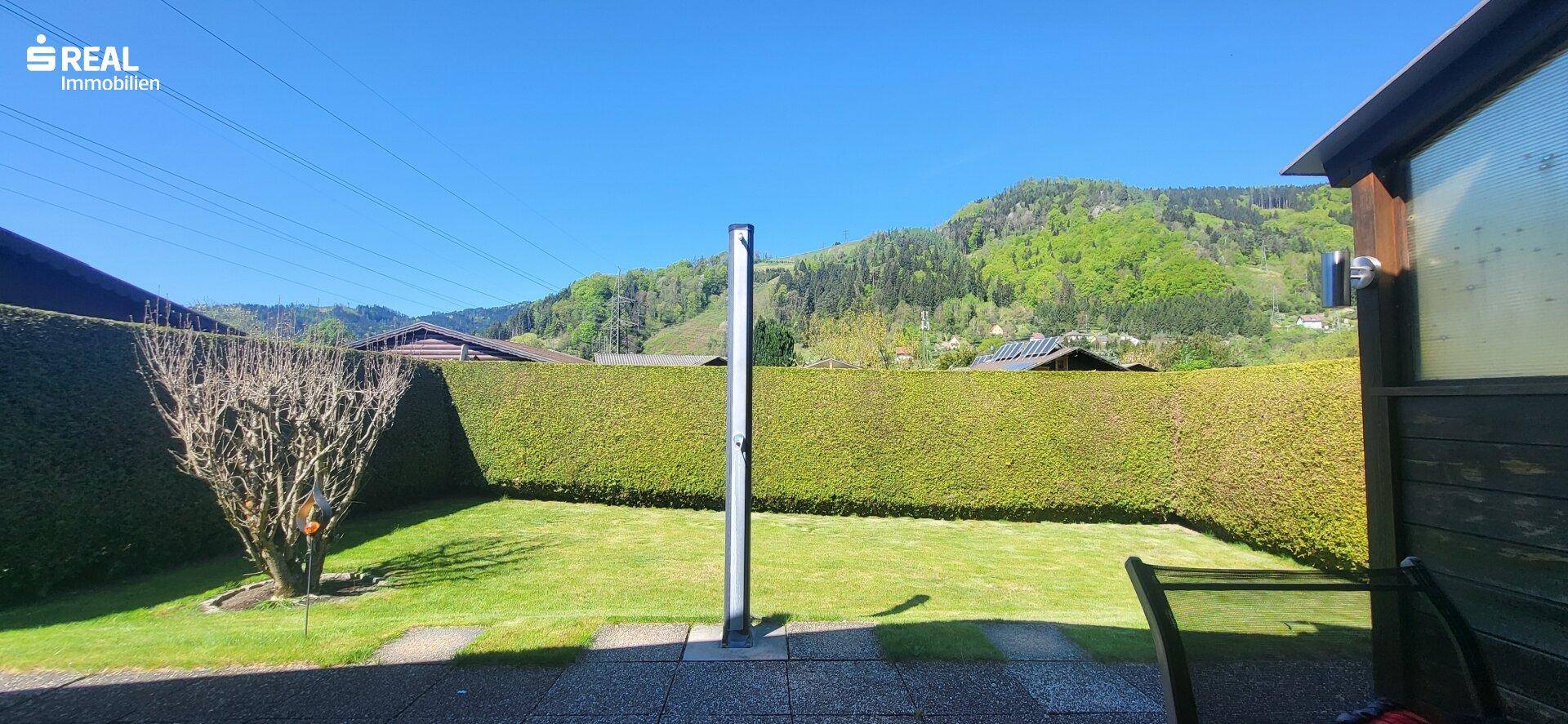 Gepflegter Garten mit Rasenfläche, hoher Hecke und Blick auf die umliegenden grünen Hügel.