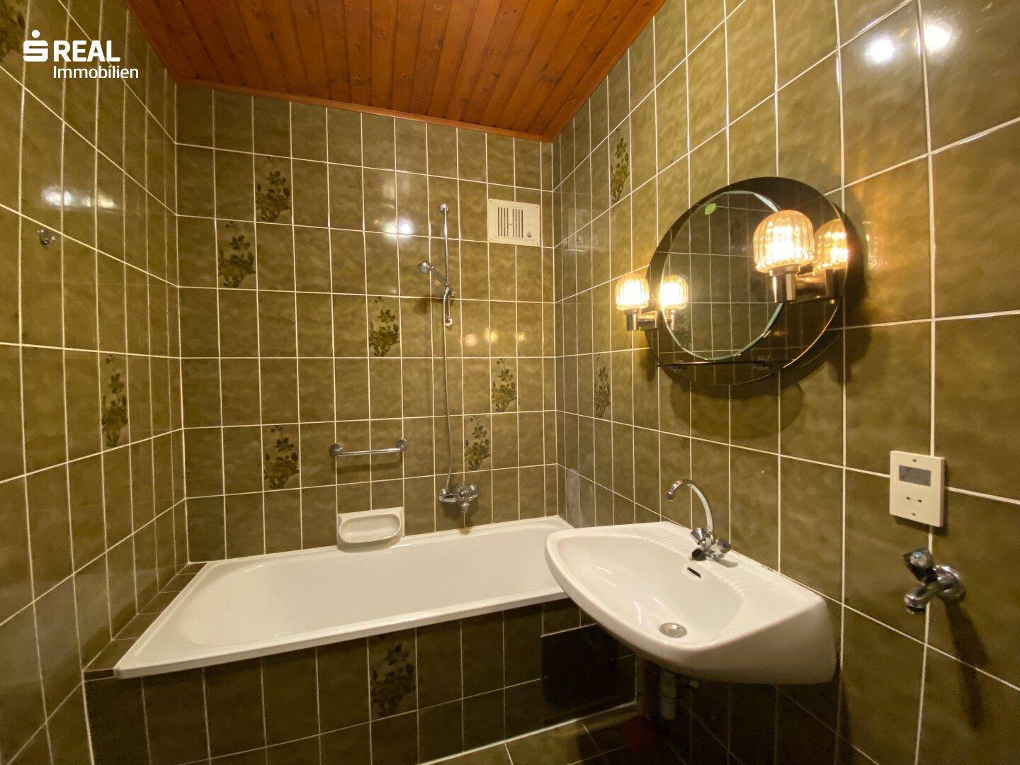 Badezimmer mit grünen Fliesen, Badewanne, Waschbecken und Holzdecke im 80er-Jahre-Stil.