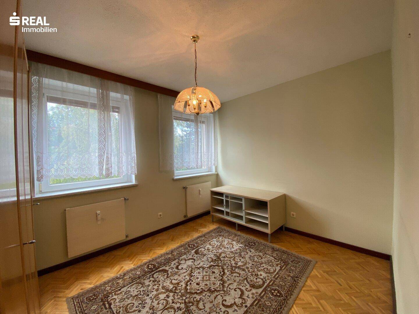 Heller Raum mit Parkettboden, zwei Fenstern und einem Teppich in der Mitte.