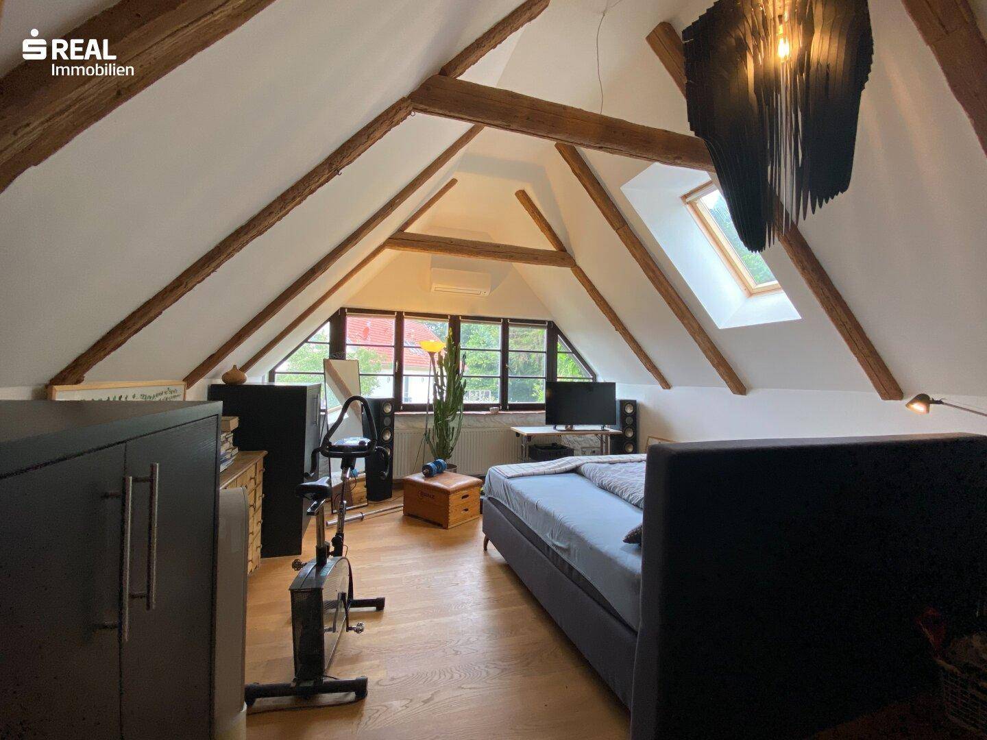 Helles Schlafzimmer im Dachgeschoss mit Holzbalken, Dachfenster und Fitnessbereich.
