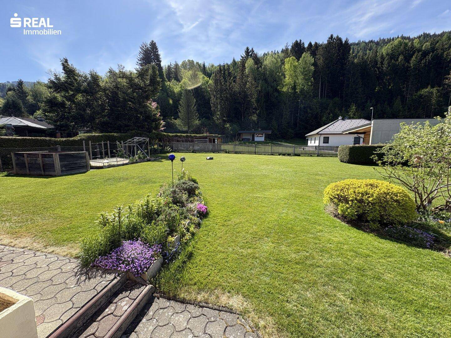 Großer, gepflegter Garten mit Blumenbeeten und weitem Blick auf den angrenzenden Wald.