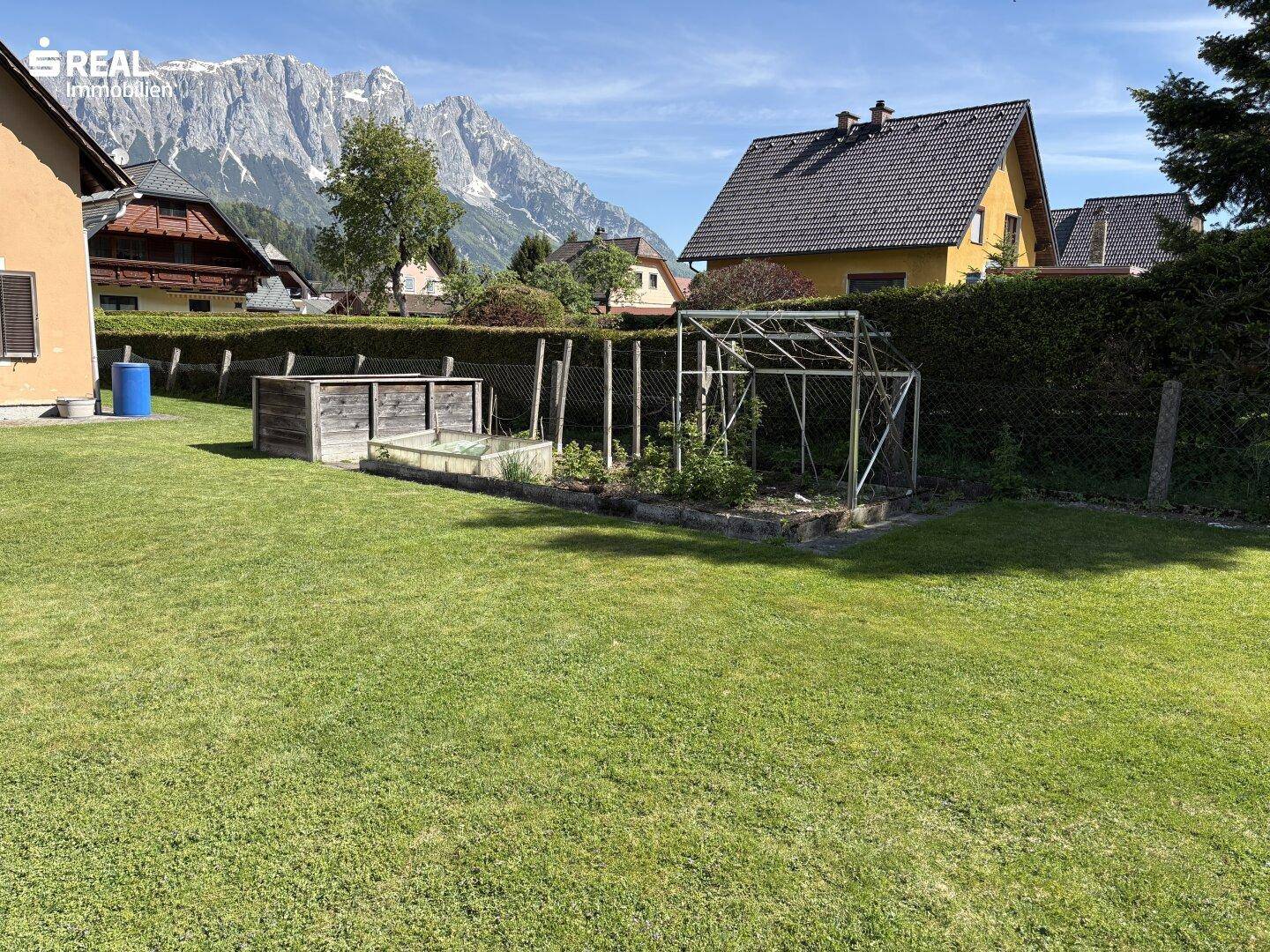 Gartenbereich mit kleinem Gewächshaus, Hochbeeten und Blick auf die umliegenden Häuser.