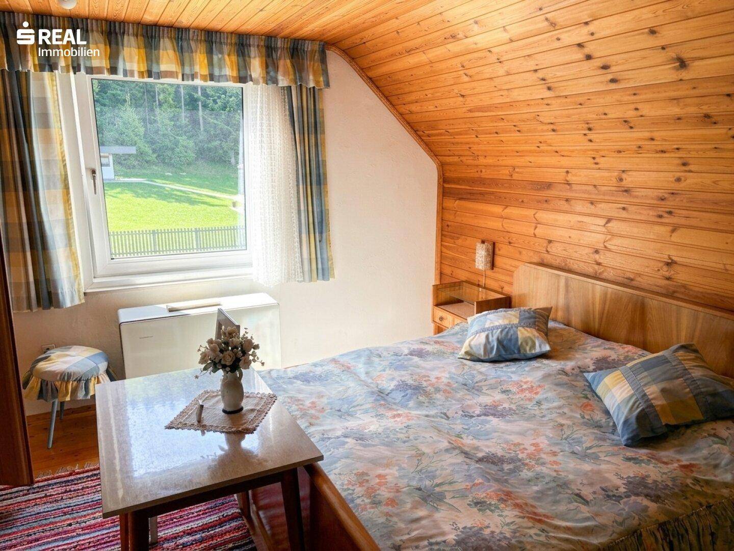 Schlafzimmer im Dachgeschoss mit Holzvertäfelung, schräger Decke und Blick ins Grüne.