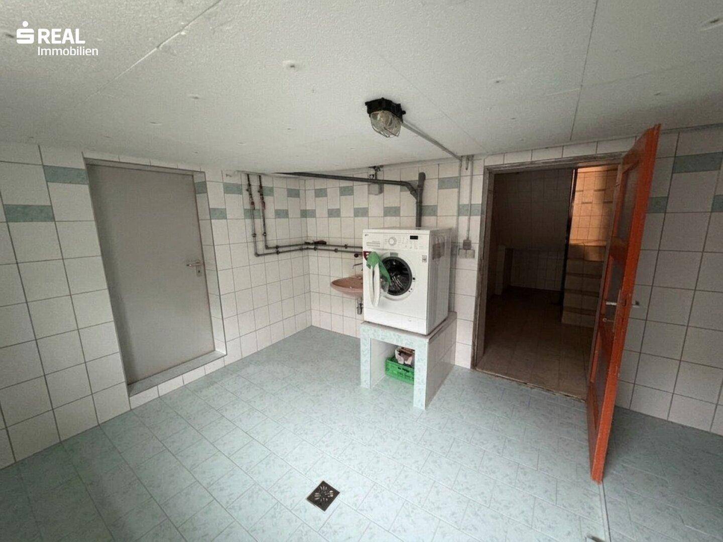 Waschküche im Keller mit Fliesenboden, Waschmaschine und Zugang zu weiteren Räumen.