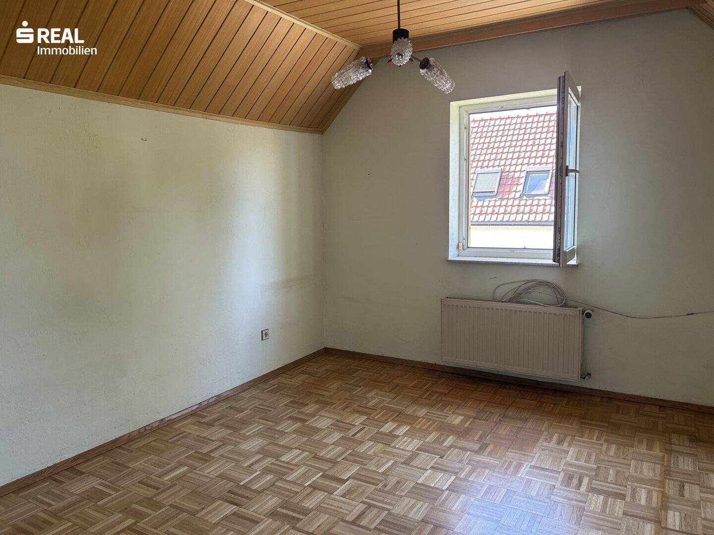 Helles Dachzimmer mit Holzdecke, Parkettboden und einem Fenster mit Blick nach draußen.