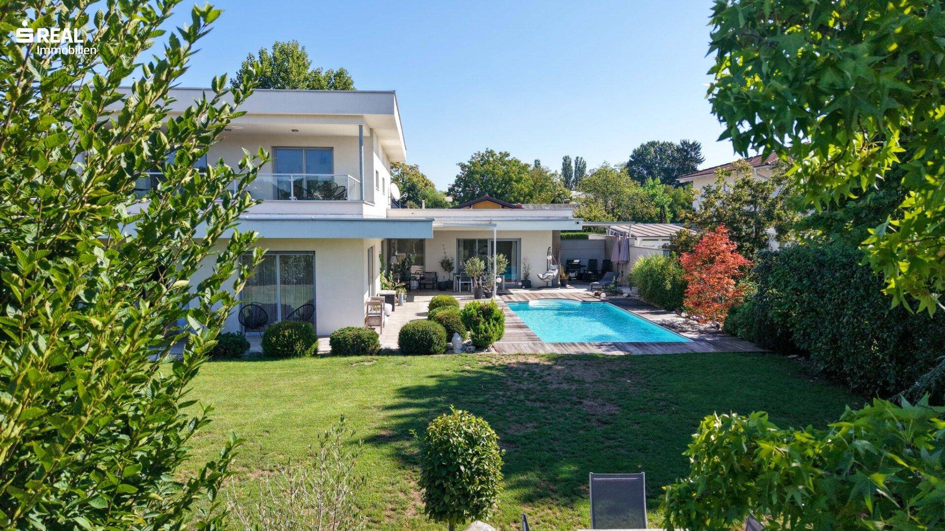 Weitläufiger Garten mit Rasenfläche, Pool und moderner Villa unter strahlend blauem Himmel.