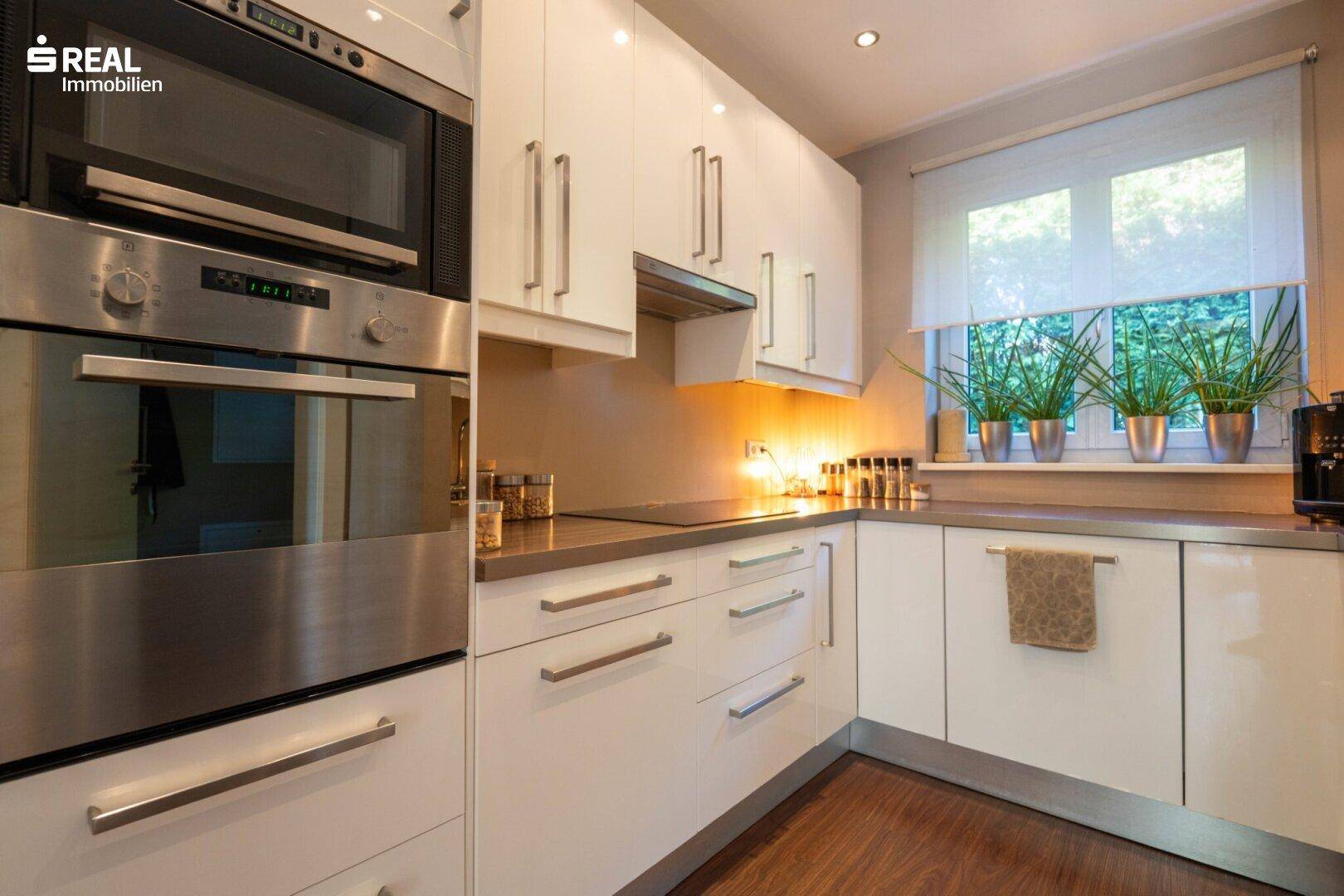 Moderne Küche mit weißen Hochglanzfronten, Edelstahlgeräten und Fenster zum Garten.