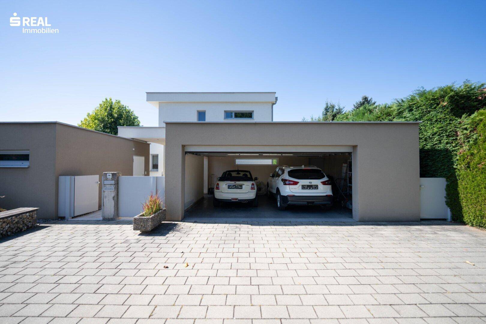 Moderne Doppelgarage mit zwei Fahrzeugen und gepflasterter Einfahrt vor dem Haus.