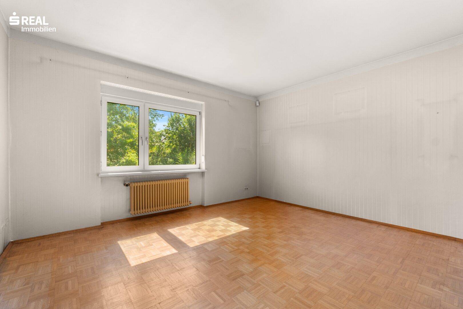 Helles Zimmer mit Fischgrätparkettboden und großem Fenster mit Blick ins Grüne.