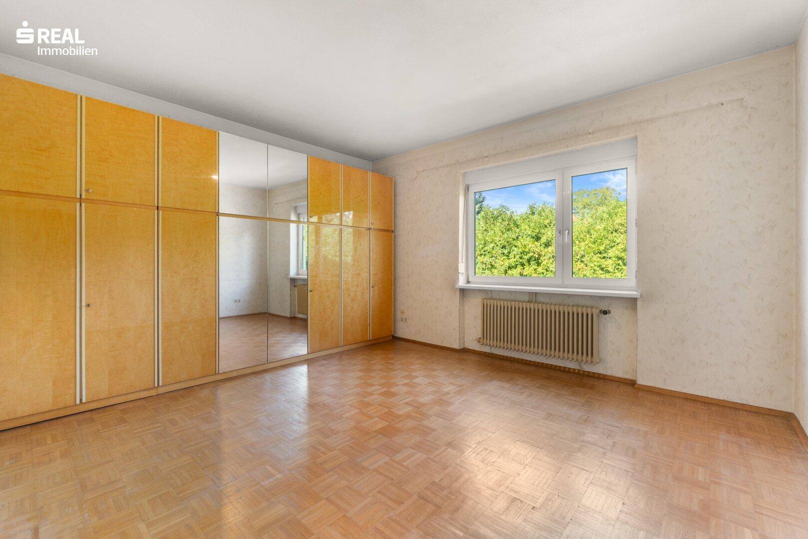 Geräumiges Schlafzimmer mit Parkettboden, Einbauschränken und Fenster mit Blick ins Grüne.