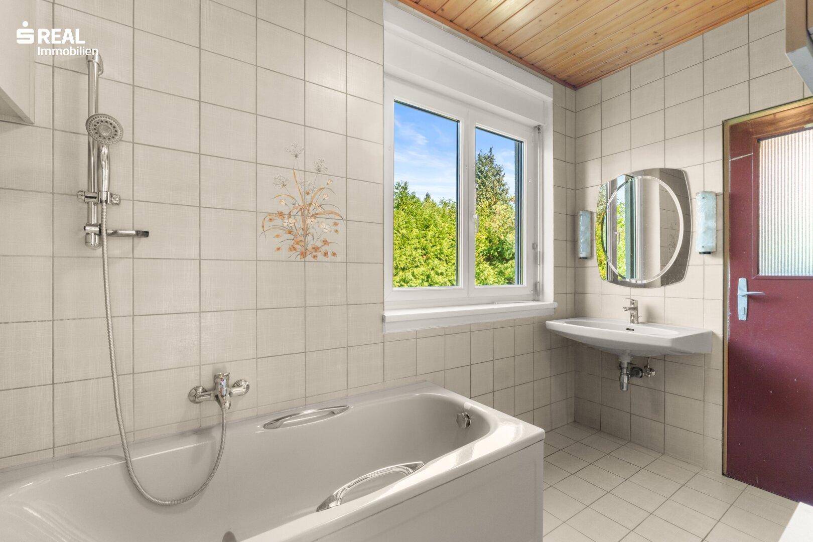 Badezimmer mit Badewanne, Waschbecken und hellen Fliesen, sowie Holzdecke.