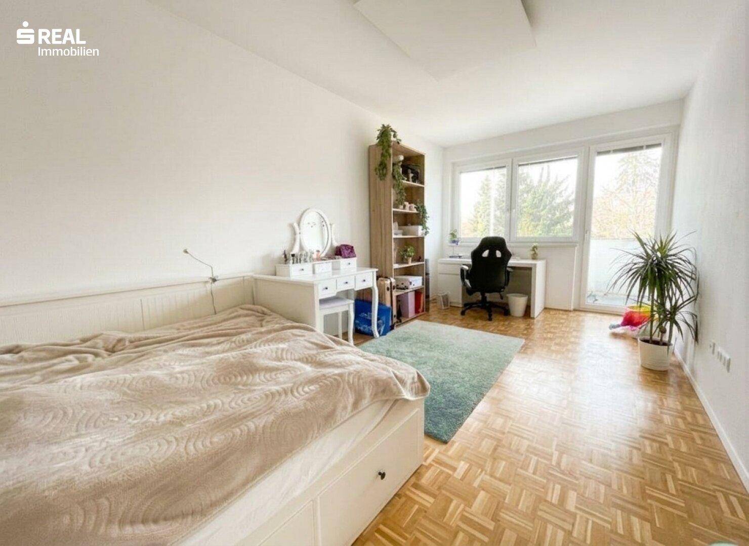 Helles Schlafzimmer mit Holzboden, großem Fenster und integriertem Arbeitsbereich.