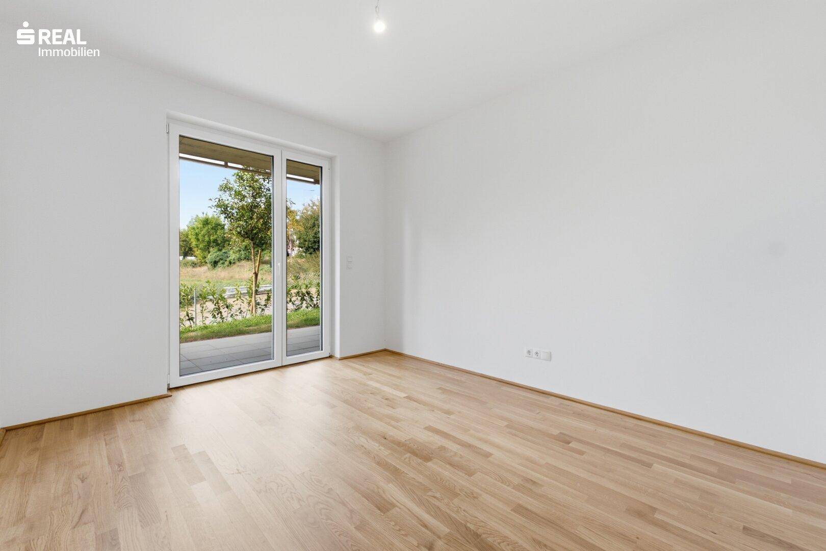 Leerer Raum mit Holzboden und großer Fenstertür, die viel Tageslicht hereinlässt.