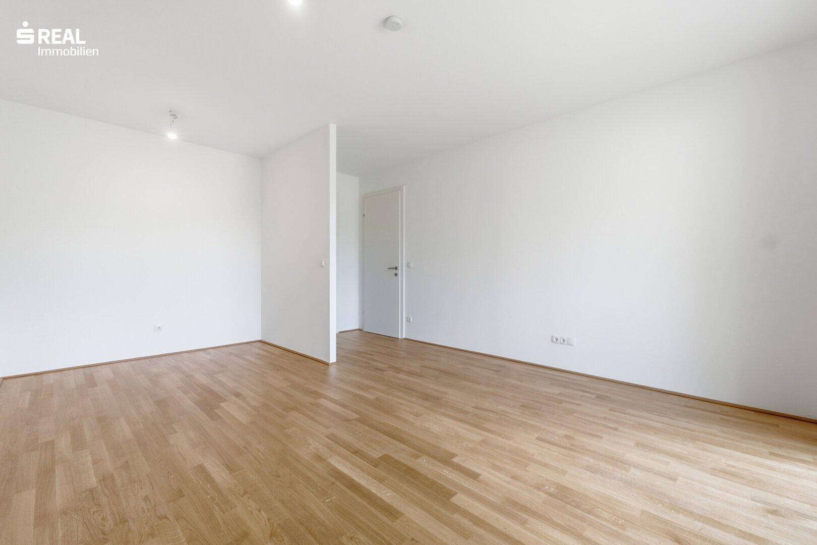 Leerer Innenraum mit Holzboden und einer Tür, die zu weiteren Räumen führt.