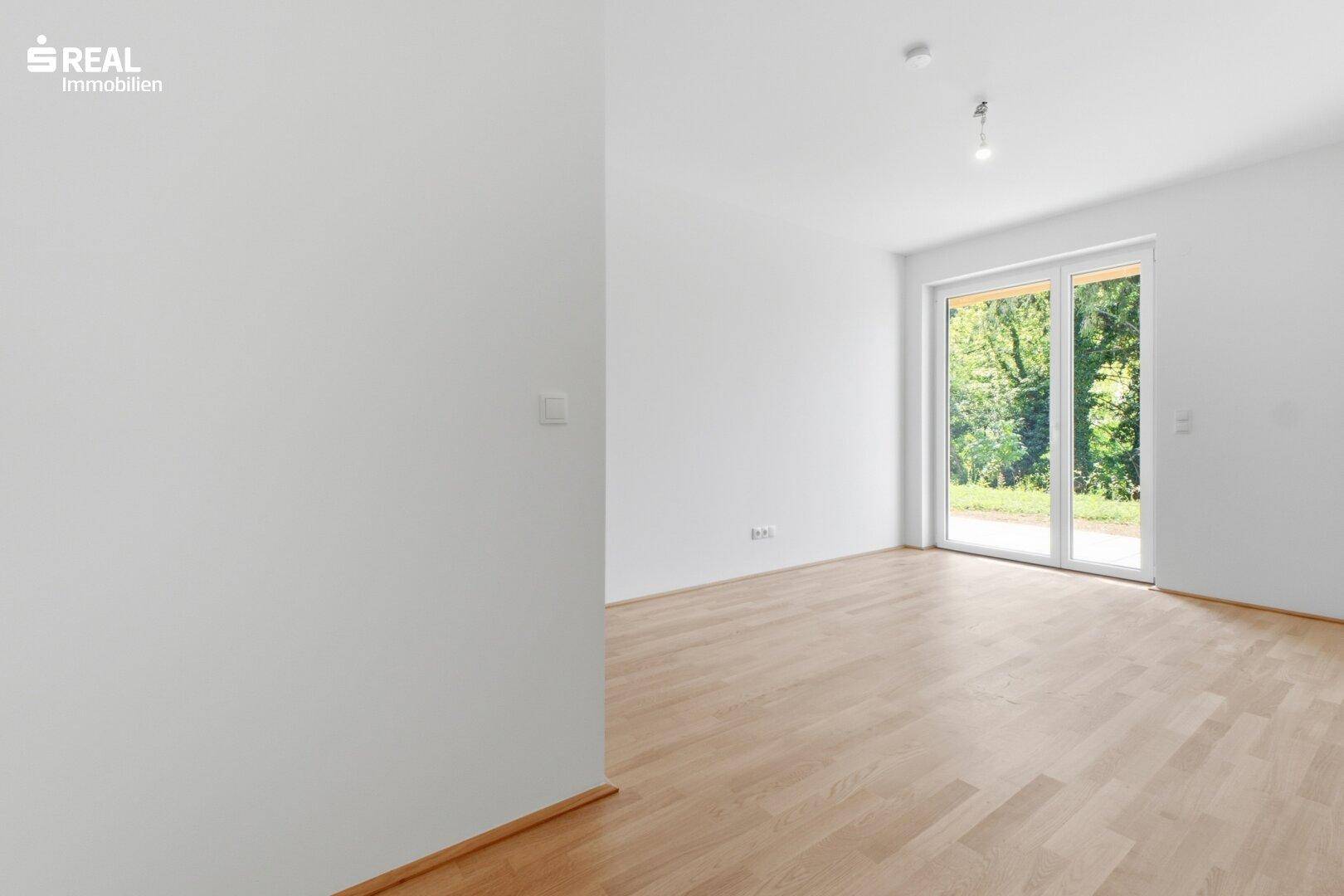 Geräumiger, heller Raum mit Holzboden und großen Fenstern, die den Blick ins Grüne freigeben.