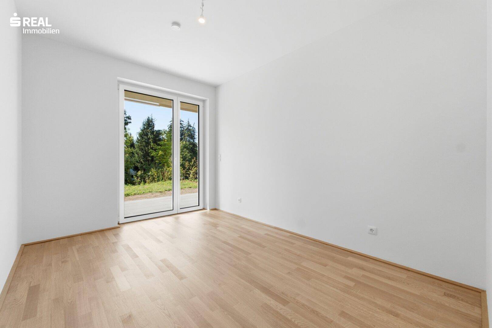 Helles Zimmer mit Holzboden und breiter Fensterfront, die viel Tageslicht und Grünblick bietet.