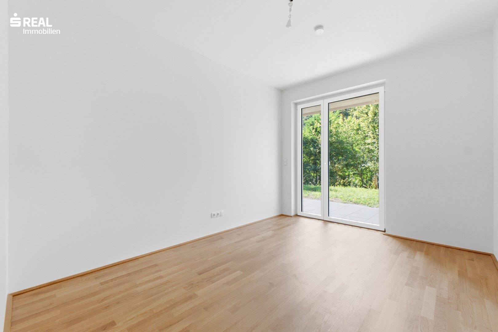 Leeres Zimmer mit Holzboden und großem Fenster, das einen direkten Zugang zum Außenbereich ermöglicht.